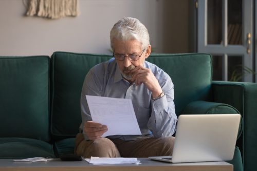 Serious mature man wearing glasses sitting on couch in living room manage budget received invoice analyzes month expenses