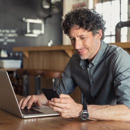 Happy mature business man sitting at cafeteria with laptop and smartphone. Businessman texting on smart phone while sitting in a pub restaurant. Portrait of senior formal man working and checking email on computer.