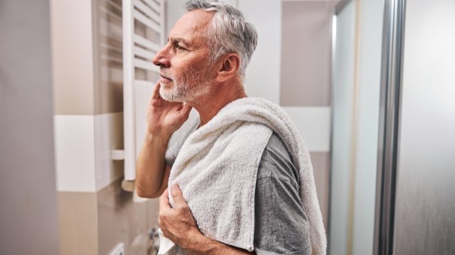 Side-view photo of a serious aged man looking in the mirror while standing in the bathroom with a towel