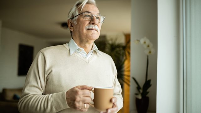 A senior man drinking a cup of coffee or tea from a mug while looking out the window