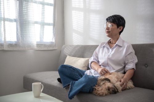 older woman petting dog on the couch next to her while looking out the window