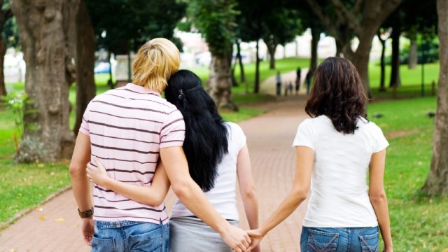 man with his arm around a woman in a park but he's holding hands with another woman behind her back