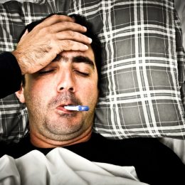 man with thermometer in his mouth feeling his forehead with a pillow and blanket
