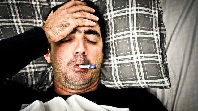 man with thermometer in his mouth feeling his forehead with a pillow and blanket