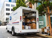 USPS delivery van stopped in front of a UPS location, unloading Amazon packages