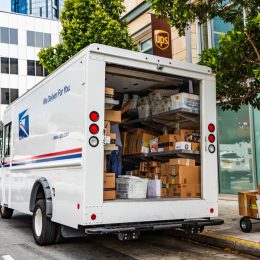 USPS delivery van stopped in front of a UPS location, unloading Amazon packages