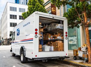 USPS delivery van stopped in front of a UPS location, unloading Amazon packages