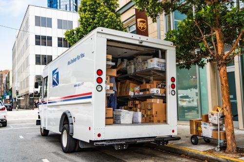 USPS delivery van stopped in front of a UPS location, unloading Amazon packages