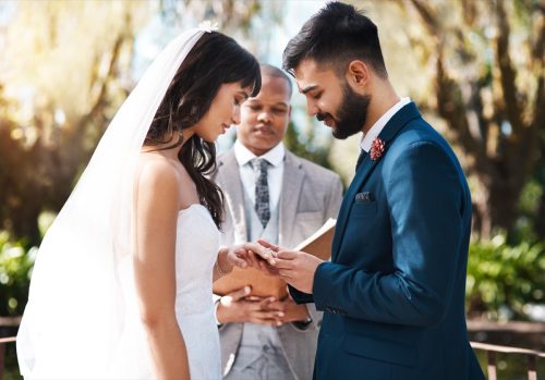Изрязан кадър на нежен млад младоженец, който нахлузва пръстен върху пръста на булката си, докато стои пред олтара в сватбения им ден