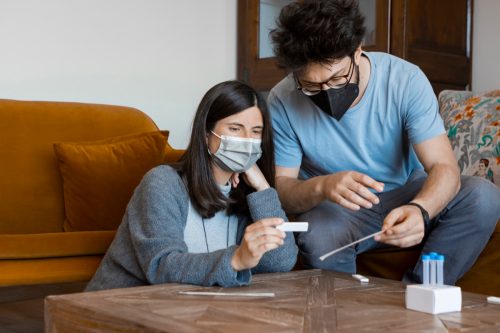 Мъж и жена на 30-те, носещи хирургически маски за лице, седят в хола вкъщи и проверяват резултатите от самоизтриващия се антигенен домашен тест за диагностика на коронавирус.