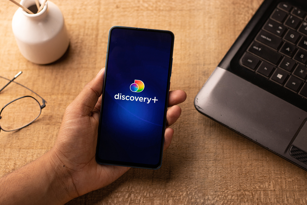 A hand holding a phone with the Discovery+ app logo