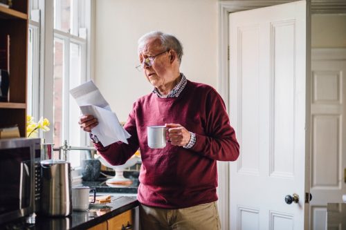 Senior man is standing in the kitchen of his home with bills in one hand