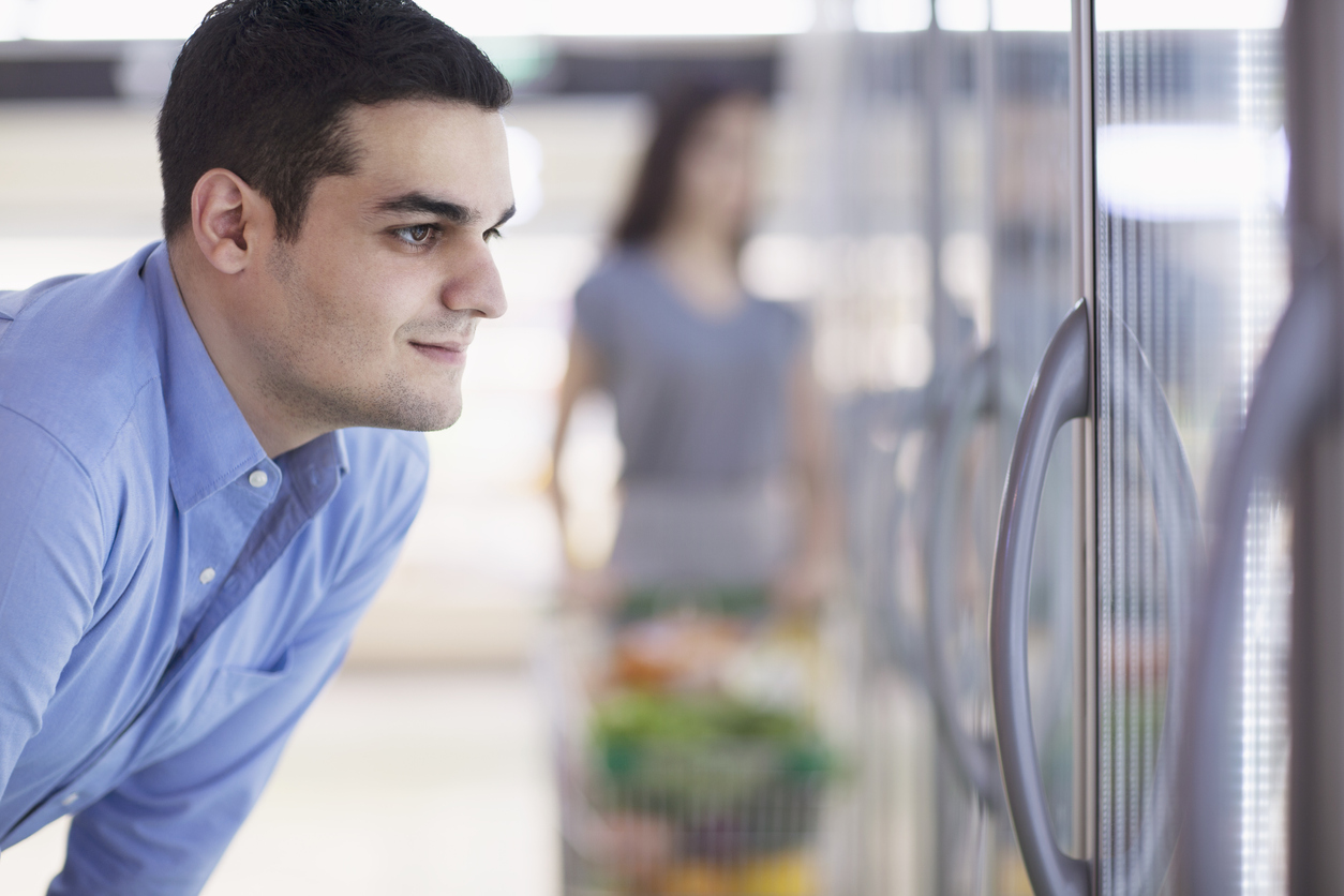 A man looking through a cooler or freezer door while shopping in the refrigerated or frozen food section in a store