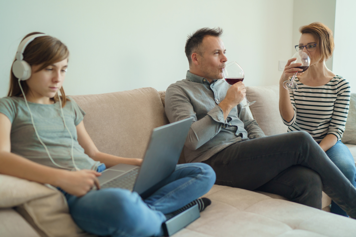 young girl sitting on couch using laptop and headphones while parents drink wine.