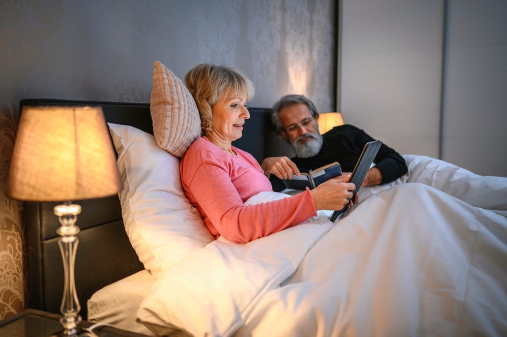 A senior couple lying in bed while using a tablet and reading before going to sleep