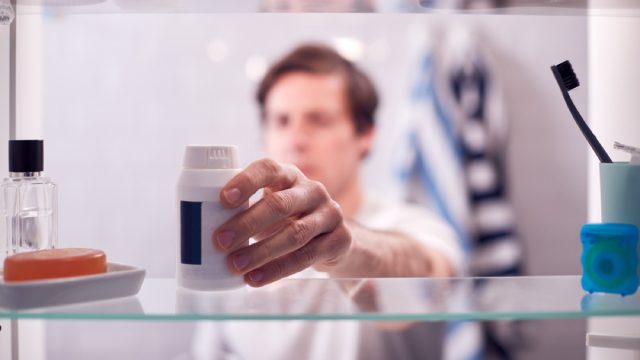 man reaching for bottle of pills from the medicine cabinet, seen from behind bathroom mirror