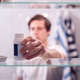 man reaching for bottle of pills from the medicine cabinet, seen from behind bathroom mirror