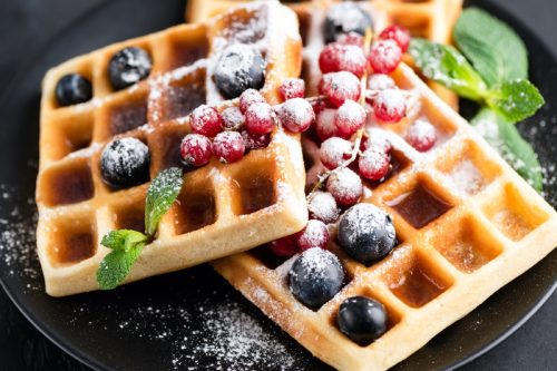 close up of square waffles with mint blueberries and berries on top along with maple syrup and powdered sugar