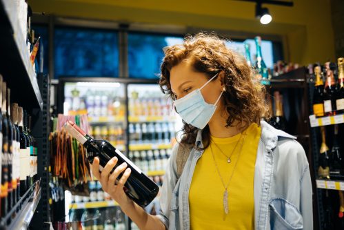 Млада жена, носеща защитна маска за лице, избира вино в магазин за хранителни стоки