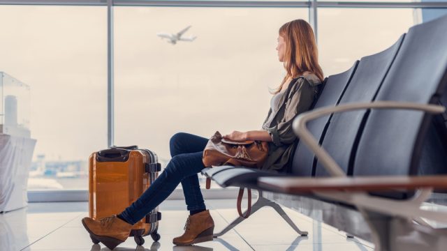 woman at airport with suitcase at her feet looking out the window at planes departing
