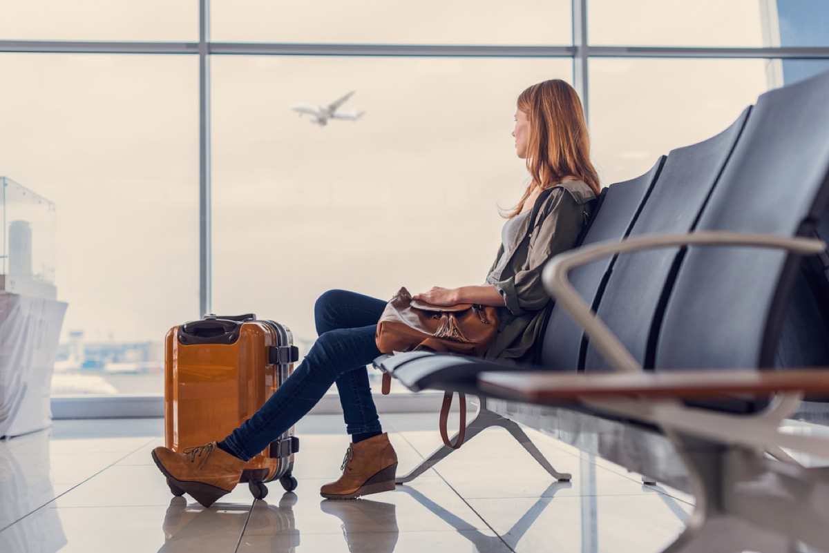 woman at airport with suitcase at her feet looking out the window at planes departing