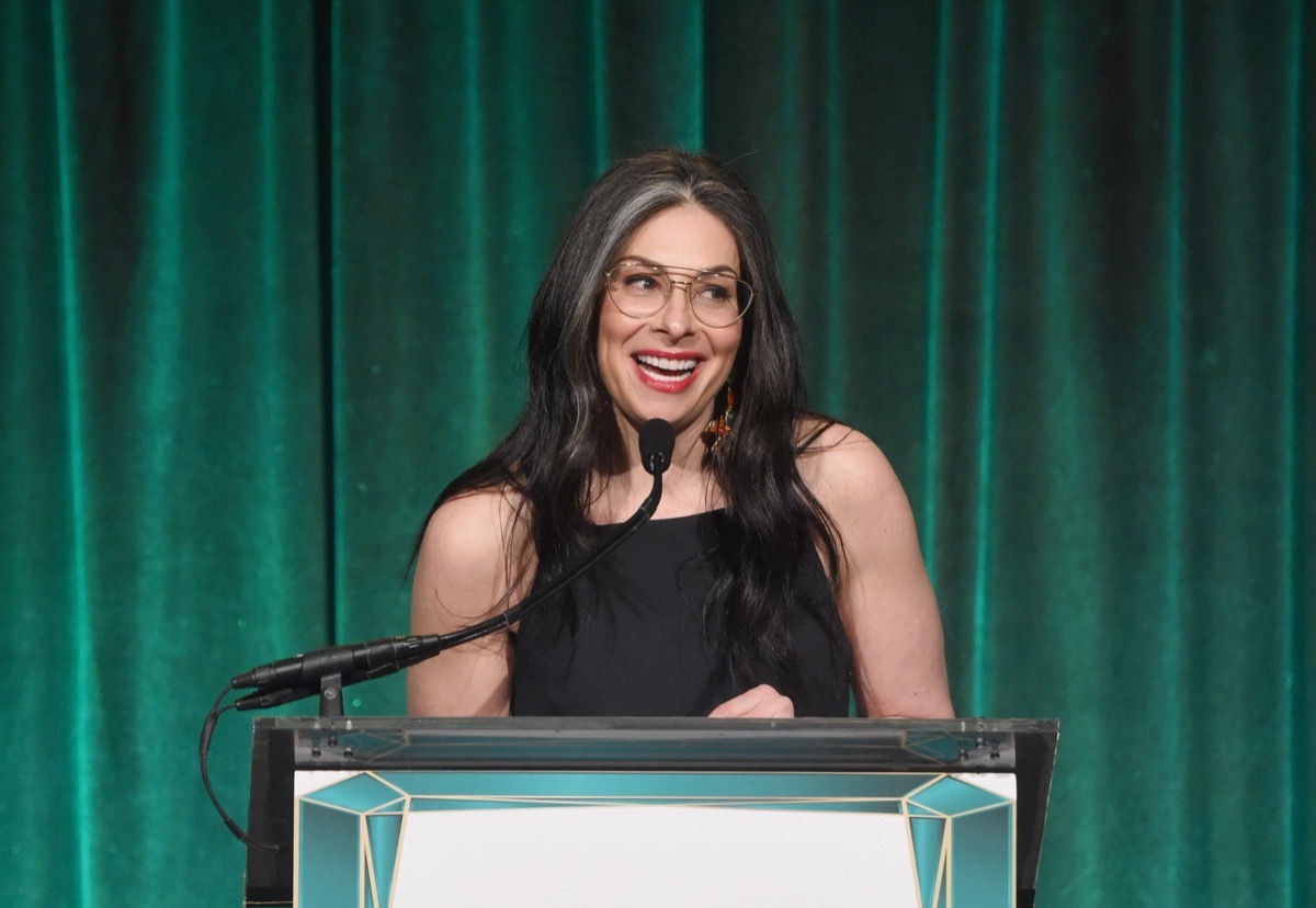 stacy london speaking at a podium with a green curtain behind her