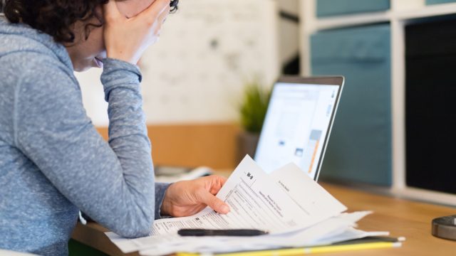 A young woman sits at a desk late at night and tries to work on her taxes. She looks discouraged as she reads a W-9 and other paperwork. Her laptop is open in the background.