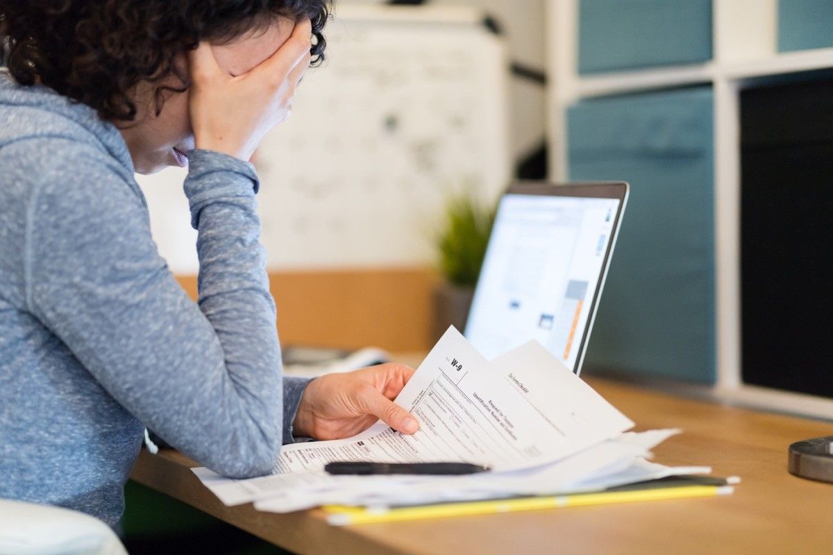 A young woman sits at a desk late at night and tries to work on her taxes. She looks discouraged as she reads a W-9 and other paperwork. Her laptop is open in the background.