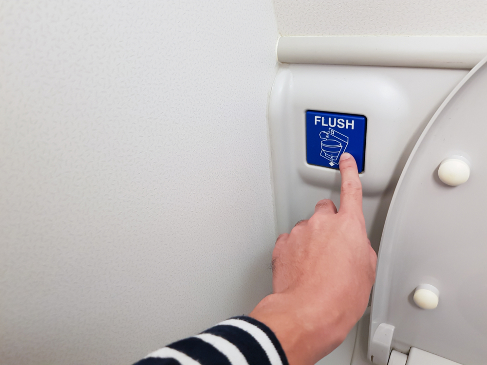 A close up of a hand pressing the flush button on the toilet in an airplane bathroom