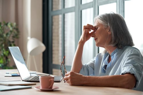 elderly white woman looking exhausted while rubbing her eyes in front of her computer
