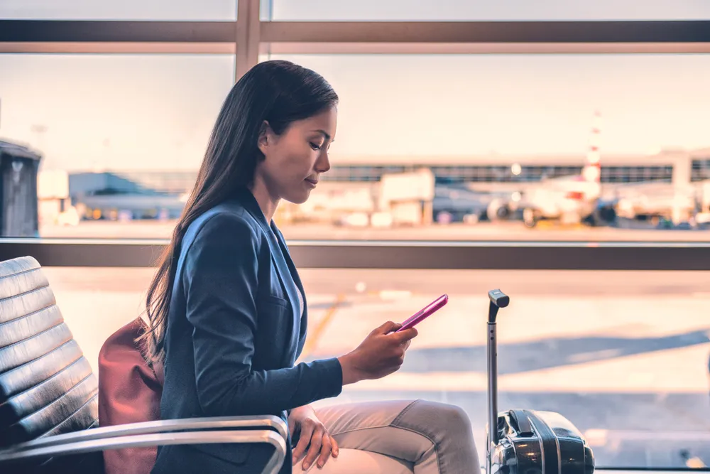 ผู้หญิงใช้โทรศัพท์ขณะนั่งอยู่ในอาคารผู้โดยสารของสนามบินเพื่อรอขึ้นเครื่อง