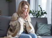 A woman blowing her nose while sick on the couch with COVID symptoms