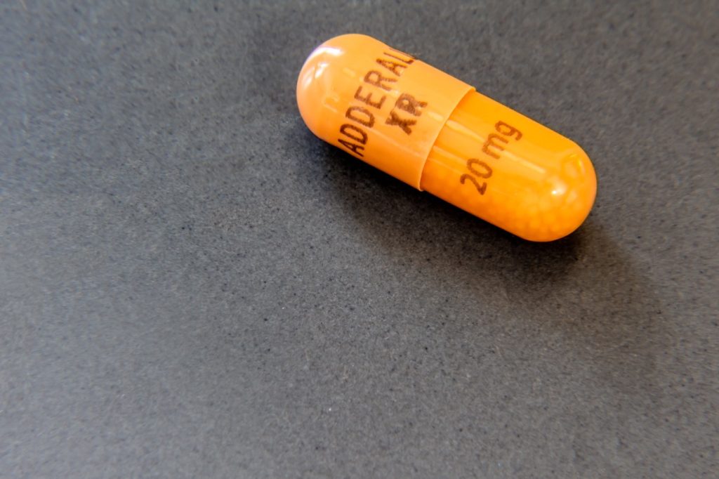 Single 20mg capsule of Adderall XR, a mixed salt amphetamine stimulant drug used in psychiatric medicine to treat ADD, ADHD and narcolepsy, on a gray surface.