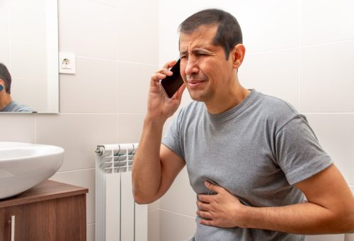 Cropped shot of an man sitting on the toilet in a bathroom suffering from stomach cramps and calling medical assistance on phone
