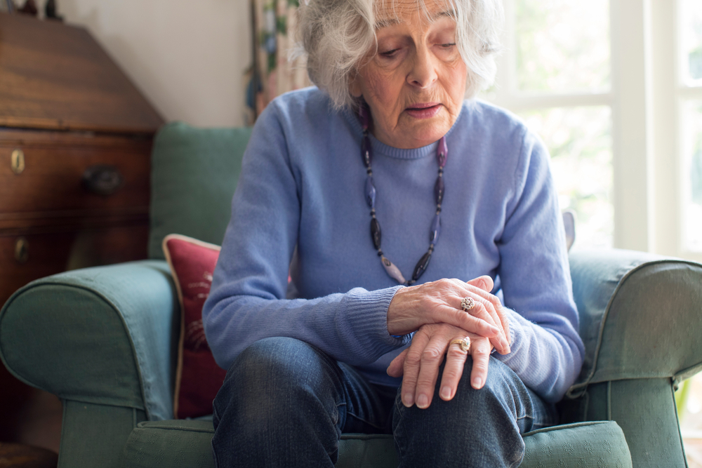 A senior woman sitting in a chair holding her hand, noticing symptoms of Parkinson's disease