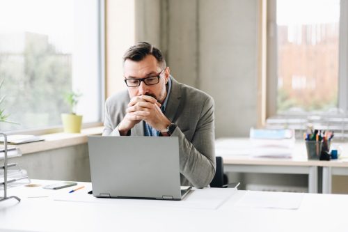 Om de afaceri chipeș de vârstă mijlocie, gânditor, într-un tricou, care lucrează la un laptop la birou.  om care lucrează la birou