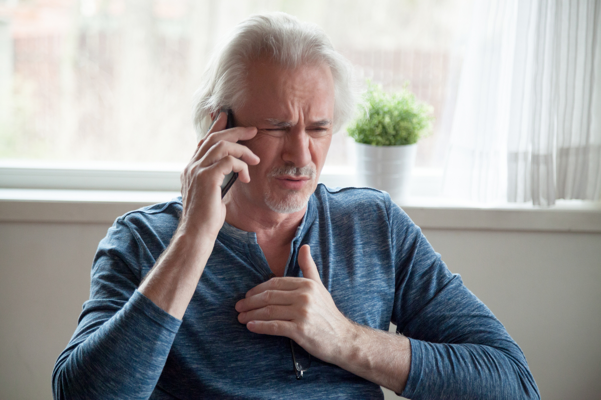 senior white man with long hair clutching his chest and talking on the phone looking worried