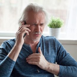 older white man with long hair clutching chest and talking on phone, looking worried