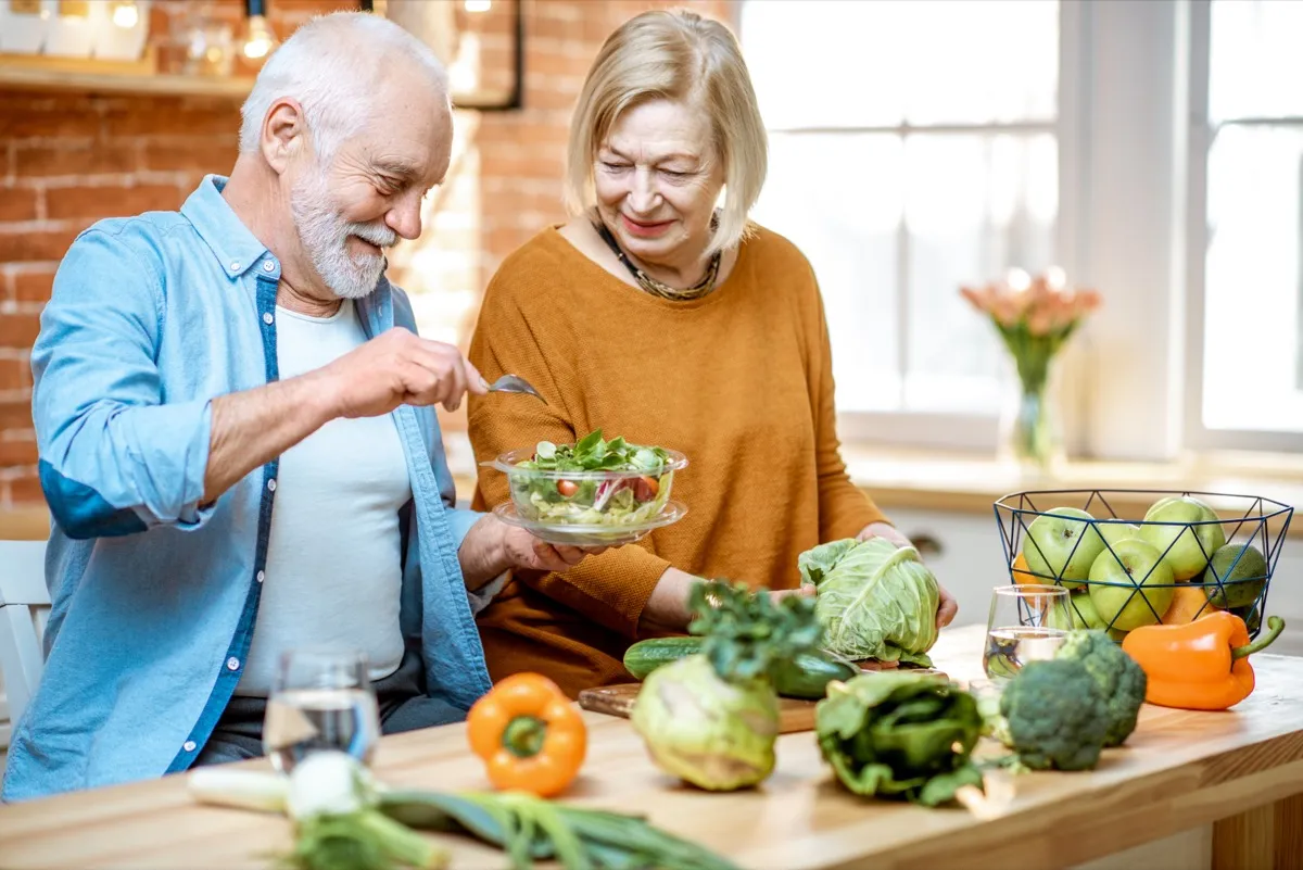 Cheerful senior couple eating salad standing together with healthy food on the kitchen at home