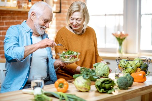 Cặp vợ chồng già vui vẻ ăn salad đứng cùng với thức ăn tốt cho sức khỏe trong bếp ở nhà