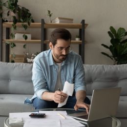 man holding a piece of paper with papers next to him while using his laptop on the couch to do taxes