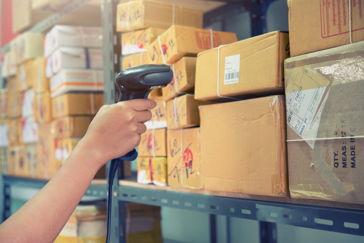 Postman worker scanning package with barcode scanner in warehouse for delivery.