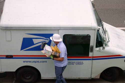 Ein Kurier des United States Postal Service trägt eine Maske und Handschuhe, während er während der COVID-19-Coronavirus-Pandemie eine Sendung von Paketen von einem Postlastwagen trägt.