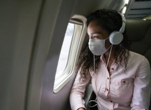 A young woman wearing a mask and headphones looking out the window of an airplane