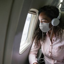 A young woman wearing a mask and headphones looking out the window of an airplane