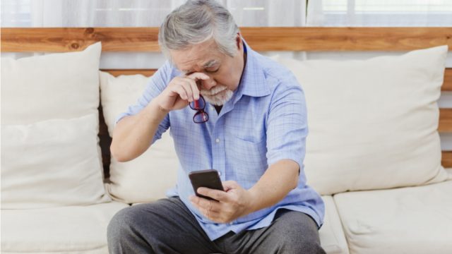 man sitting on couch holding phone outstretched and rubbing his eyes
