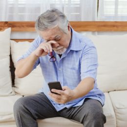 man sitting on couch holding phone outstretched and rubbing his eyes