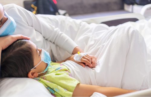 Eine junge Mutter, die eine schützende Gesichtsmaske trägt, hält die Hand ihres Sohnes, während sie mit ihm auf einem Krankenhausbett liegt.  Ein emotionaler Familienmoment.  Zugang zu den Blutgefäßen des Babys