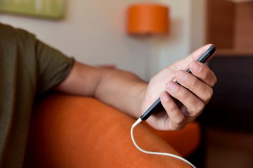closeup of a young man using a smpartphone with a cable plugged into it, sitting in a comfortable sofa indoors