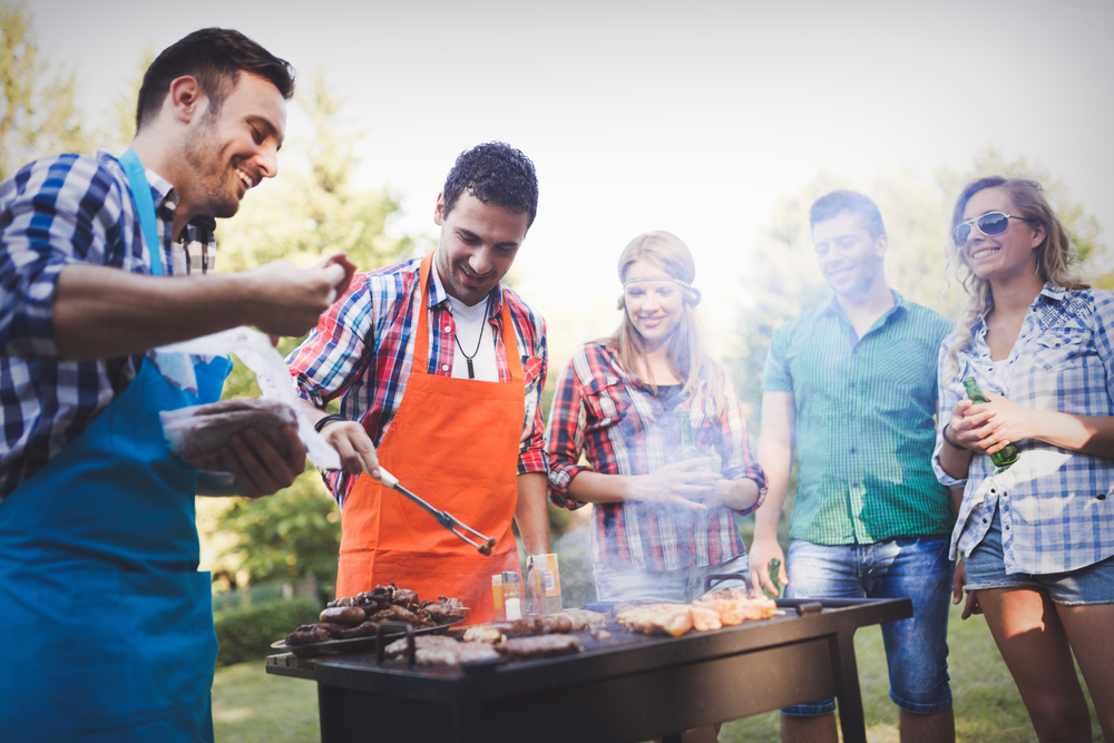 A group of friends standing around a barbecue while meat is grilled